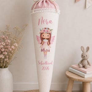 May include: A conical gift cone featuring a light pink fabric top with a rope tie and white lace trim. The off-white body displays a fairy illustration, the name Nora, and the text Schulkind 2026 in pink script, centered on the front surface.