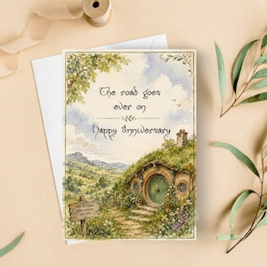 May include: A rectangular greeting card featuring a watercolour illustration of a green-roofed hobbit hole nestled in a grassy hill. The text reads: The road goes ever on Happy Anniversary. A small wooden sign in the foreground says: not all those who wander are lost.