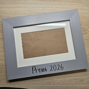 May include: A rectangular picture frame with a light grey wood grain finish and a white mat board. The bottom edge of the frame features the black text Prom 2026. The centre of the frame contains a brown backing board with visible metal fasteners.
