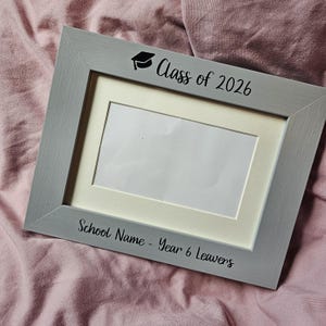 May include: A rectangular grey picture frame with a white mat board. The top of the frame features a black graduation cap icon and the text "Class of 2026". The bottom of the frame displays the black text "School Name - Year 6 Leavers".