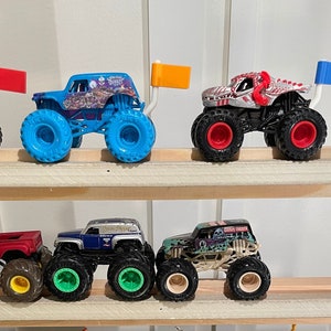 May include: A collection of monster truck toys on a wooden shelf. The trucks are various colors, including blue, red, green, and black. Each truck has a unique design and features a flag on the back.