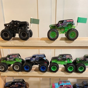 May include: A collection of monster truck toys displayed on a wooden shelf. The trucks are various colors, including green, black, orange, and pink. Some of the trucks have flags attached to the back.