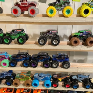 May include: A collection of monster truck toys displayed on a wooden shelf. The trucks are various colors, including green, blue, red, and black. Some of the trucks have flags attached to the back.