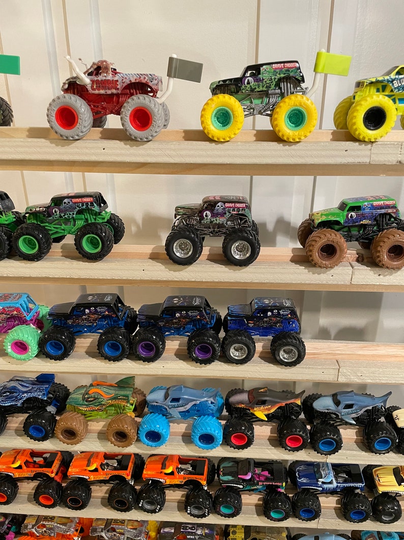 May include: A collection of monster truck toys displayed on a wooden shelf. The trucks are various colors, including green, blue, red, and black. Some of the trucks have flags attached to the back.