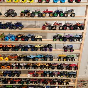 May include: A collection of monster truck toys displayed on a wooden shelf. The trucks are various colors, including red, blue, green, yellow, and black. Some of the trucks have flames painted on them, while others have different designs. The trucks are all on wheels and have different designs.