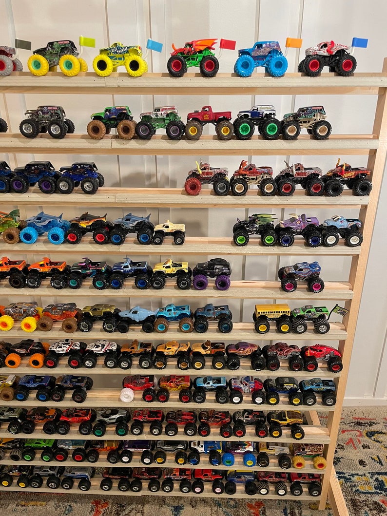 May include: A collection of monster truck toys displayed on a wooden shelf. The trucks are various colors, including red, blue, green, yellow, and black. Some of the trucks have flames painted on them, while others have different designs. The trucks are all on wheels and have different designs.
