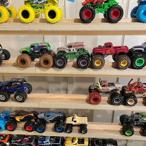May include: A collection of monster trucks displayed on a wooden shelf. The trucks are various colors, including green, red, blue, yellow, and black. Some of the trucks have flags attached to them.
