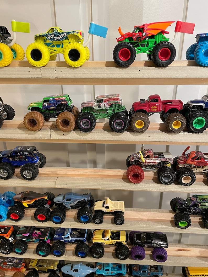 May include: A collection of monster trucks displayed on a wooden shelf. The trucks are various colors, including green, red, blue, yellow, and black. Some of the trucks have flags attached to them.