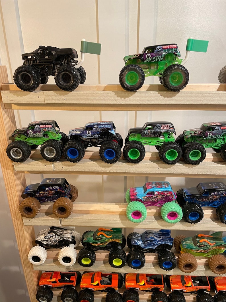 May include: A collection of monster truck toys displayed on a wooden shelf. The trucks are various colors, including green, black, orange, and pink. Some of the trucks have flags attached to the back.