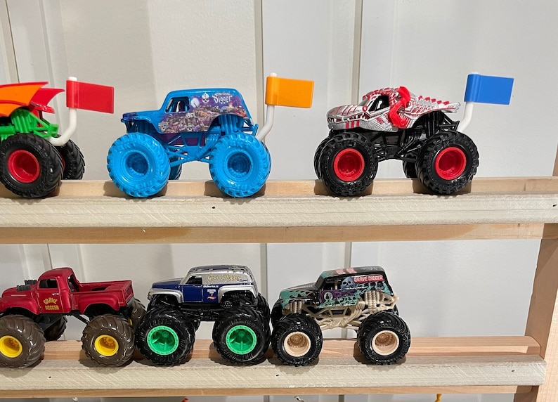 May include: A collection of monster truck toys on a wooden shelf. The trucks are various colors, including blue, red, green, and black. Each truck has a unique design and features a flag on the back.