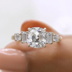Puede incluir: Un anillo de compromiso de oro con una gran piedra central de diamante de talla cojín y diamantes más pequeños de talla baguette a cada lado.