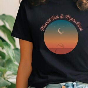Może przedstawiać: Czarny T-shirt z grafiką przedstawiającą delfina skaczącego przed rosnącym księżycem i gwiazdami. Grafika jest otoczona kółkiem z tekstem "Moonlit Tides & Mystic Rides".