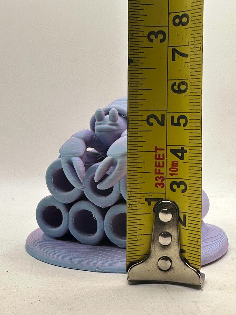 Puede incluir: Escultura impresa en 3D de un cangrejo morado claro con una base de arrecife de coral. El cangrejo mide aproximadamente 5 cm de altura. La escultura presenta un dise&ntilde;o detallado con una superficie texturizada y est&aacute; sobre un fondo liso.