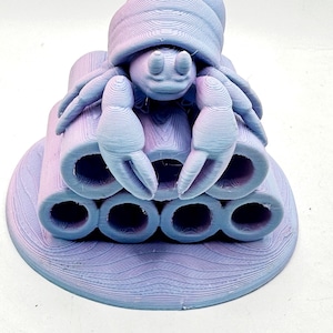 Puede incluir: Escultura impresa en 3D de un cangrejo con caparaz&oacute;n, pinzas y m&uacute;ltiples estructuras tubulares. La escultura es en tonos morados y azules, con un aspecto texturizado y en capas. El cangrejo se asienta sobre una base circular.