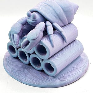 Puede incluir: Escultura impresa en 3D de un cangrejo ermita&ntilde;o en tonos lavanda y azul. El cangrejo est&aacute; posado sobre una pila de seis tubos cil&iacute;ndricos, todo sobre una base circular con patrones de olas. El cangrejo tiene pinzas y un caparaz&oacute;n detallados.