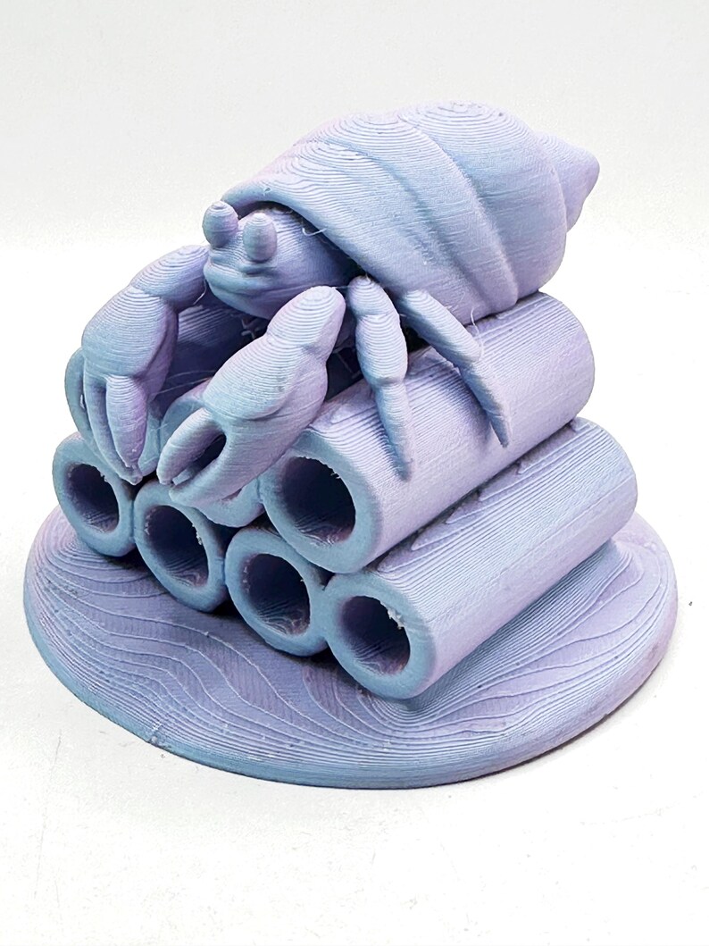 Puede incluir: Escultura impresa en 3D de un cangrejo ermita&ntilde;o en tonos lavanda y azul. El cangrejo est&aacute; posado sobre una pila de seis tubos cil&iacute;ndricos, todo sobre una base circular con patrones de olas. El cangrejo tiene pinzas y un caparaz&oacute;n detallados.