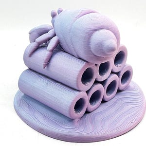 Puede incluir: Escultura impresa en 3D de un insecto estilizado con cuerpo redondeado y m&uacute;ltiples patas, posado sobre una pila de tubos cil&iacute;ndricos. La escultura es un degradado de lavanda y azul claro, con superficie texturizada y base circular.