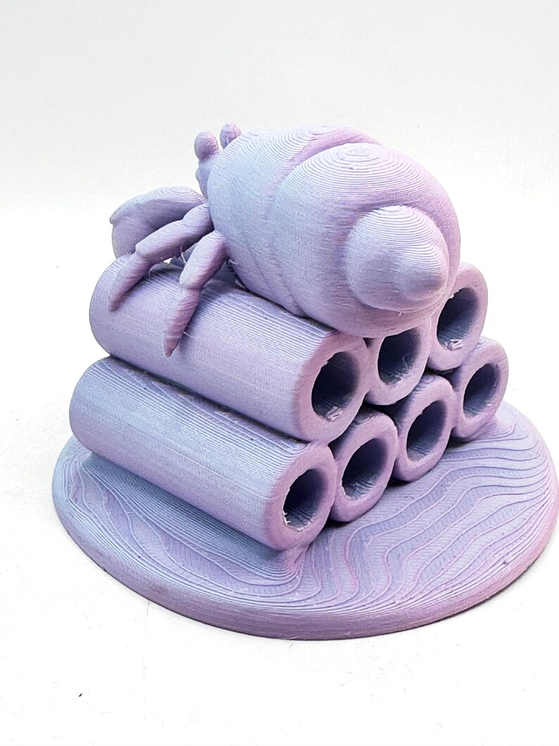 Puede incluir: Escultura impresa en 3D de un insecto estilizado con cuerpo redondeado y m&uacute;ltiples patas, posado sobre una pila de tubos cil&iacute;ndricos. La escultura es un degradado de lavanda y azul claro, con superficie texturizada y base circular.