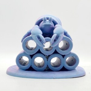 Puede incluir: Una figura de cangrejo azul claro impresa en 3D, posada sobre una pila de formas cil&iacute;ndricas. El cangrejo tiene pinzas detalladas y un caparaz&oacute;n texturizado. La base es de un azul claro a juego, creando un dise&ntilde;o cohesivo. La est&eacute;tica general es caprichosa y decorativa.