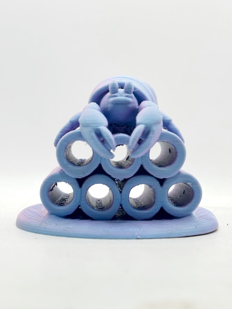 Puede incluir: Una figura de cangrejo azul claro impresa en 3D, posada sobre una pila de formas cil&iacute;ndricas. El cangrejo tiene pinzas detalladas y un caparaz&oacute;n texturizado. La base es de un azul claro a juego, creando un dise&ntilde;o cohesivo. La est&eacute;tica general es caprichosa y decorativa.