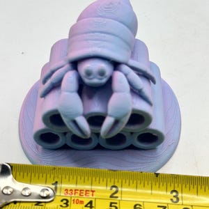 Puede incluir: Una figura de cangrejo ermita&ntilde;o impresa en 3D en tonos morados y azules. El cangrejo est&aacute; posado sobre una pila de estructuras cil&iacute;ndricas. La figura mide aproximadamente 5 cm de ancho, medido con una cinta m&eacute;trica amarilla.