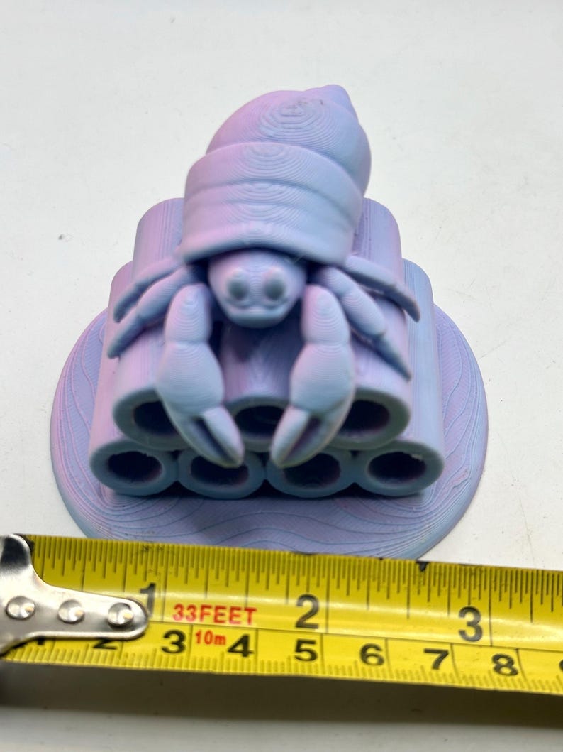 Puede incluir: Una figura de cangrejo ermita&ntilde;o impresa en 3D en tonos morados y azules. El cangrejo est&aacute; posado sobre una pila de estructuras cil&iacute;ndricas. La figura mide aproximadamente 5 cm de ancho, medido con una cinta m&eacute;trica amarilla.