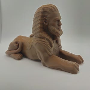 May include: A brown, 3D-printed sphinx statue. The sculpture features a human head with a beard and detailed hair, atop a lion's body. The sphinx is in a seated position, with its paws extended. The statue is made of a light brown material.