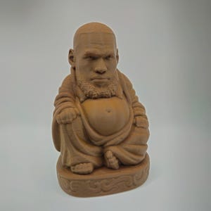 May include: A brown, 3D-printed Buddha statue with a human face, featuring a beard and a bald head. The figure is seated in a meditative pose on a decorative base. The statue is made of a light brown material.