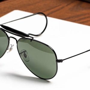 May include: Black aviator sunglasses with green lenses. The sunglasses have a black frame and a distinctive brow bar. The temples curve around the ears. The sunglasses are resting on a white surface.