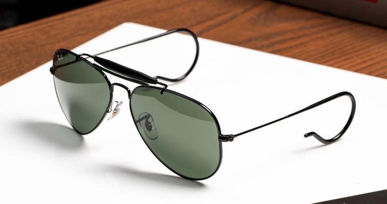 May include: Black aviator sunglasses with green lenses. The sunglasses have a black frame and a distinctive brow bar. The temples curve around the ears. The sunglasses are resting on a white surface.