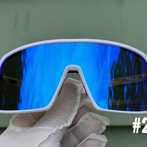May include: White-framed sunglasses with reflective blue lenses. The sunglasses have a modern, wraparound design. The number "#2" is visible in the lower right corner. The lenses reflect a bright blue hue, creating a striking contrast with the white frame.