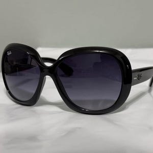 Könnte beinhalten: Schwarze übergroße Sonnenbrille mit dunklen Verlaufsgläsern. Die Sonnenbrille hat einen dicken, abgerundeten Rahmen und das Ray-Ban-Logo ist auf den Bügeln sichtbar. Die Brille ist vor einem weißen Hintergrund platziert.