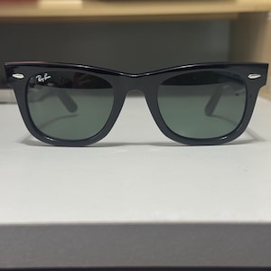 May include: Black Ray-Ban sunglasses featuring a classic wayfarer frame design with dark green tinted lenses. The frames have silver-toned metal accents on the front corners and the Ray-Ban logo is printed in white on the upper corner of the left lens.