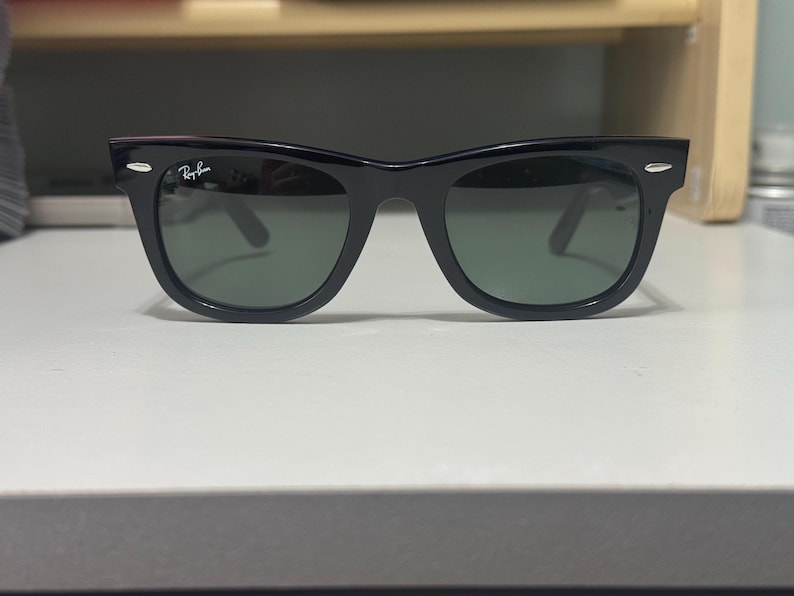 May include: Black Ray-Ban sunglasses featuring a classic wayfarer frame design with dark green tinted lenses. The frames have silver-toned metal accents on the front corners and the Ray-Ban logo is printed in white on the upper corner of the left lens.