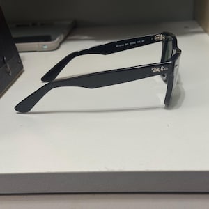 May include: Black Ray-Ban sunglasses viewed from the side, featuring a classic thick frame design. The temple arm displays the Ray-Ban logo in white lettering near the hinge, with model information printed on the inner side of the temple arm.