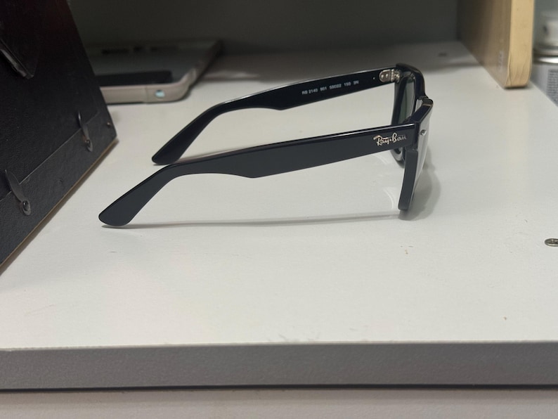 May include: Black Ray-Ban sunglasses viewed from the side, featuring a classic thick frame design. The temple arm displays the Ray-Ban logo in white lettering near the hinge, with model information printed on the inner side of the temple arm.