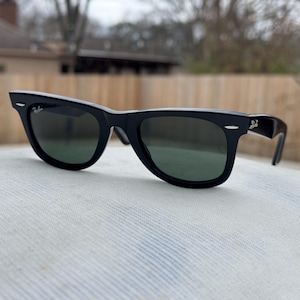 May include: Black Ray-Ban Wayfarer sunglasses featuring a thick plastic frame, dark green tinted lenses, and silver-tone accents on the front corners. The brand logo is printed in white on the upper right lens and on the outer side of the temple.