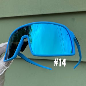 May include: Bright blue sport sunglasses with mirrored lenses. The sunglasses have a wraparound design and a bold, modern aesthetic. The number "#14" is printed in white on the lower right side of the frame. The lenses reflect the surrounding environment.