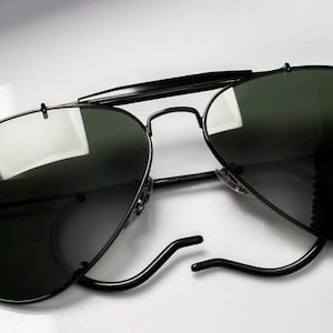 May include: Black aviator sunglasses with dark green lenses. The sunglasses have a black metal frame and the Ray-Ban logo is visible on the left lens. The sunglasses are resting on a white surface.