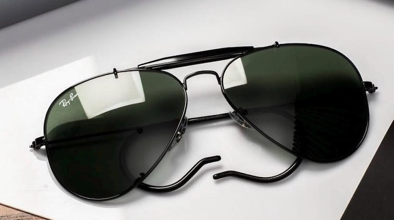 May include: Black aviator sunglasses with dark green lenses. The sunglasses have a black metal frame and the Ray-Ban logo is visible on the left lens. The sunglasses are resting on a white surface.