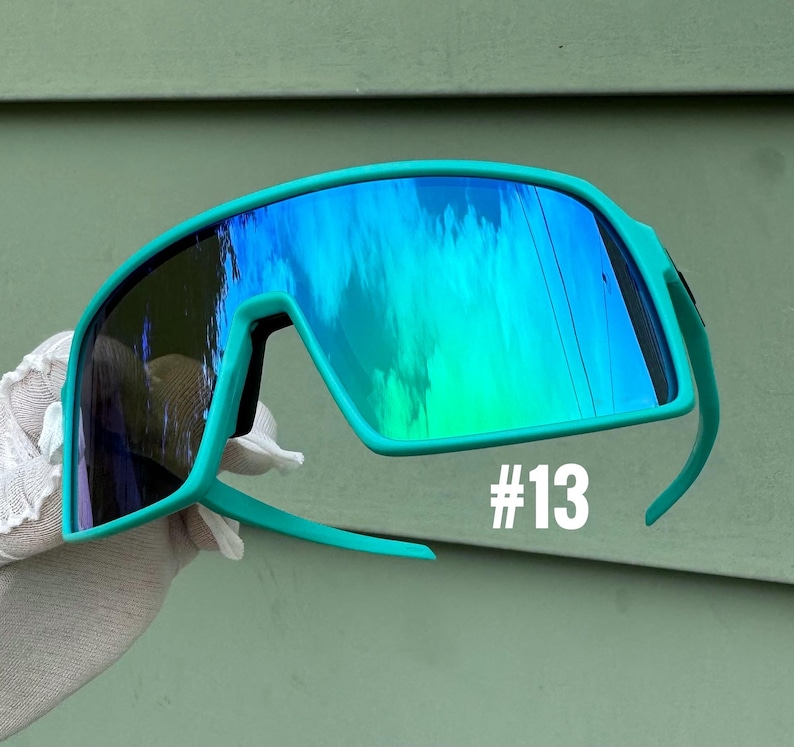 May include: Teal-colored sports sunglasses with mirrored lenses reflecting blue and green hues. The sunglasses have a thick frame and curved temples. The number "#13" is printed in white on the lower right side of the frame.