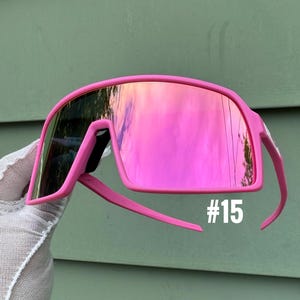 May include: Bright pink sunglasses with mirrored lenses. The sunglasses have a bold, wraparound design with a thick frame and arms. The number "#15" is printed in white on the lower right side of the frame.