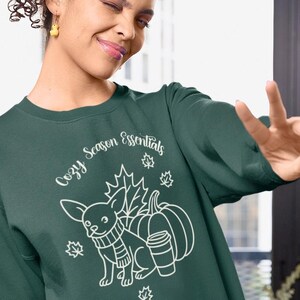 Peut inclure: Un sweat-shirt vert foncé avec le texte "Cozy Season Essentials". Le motif présente un chihuahua portant une écharpe, une citrouille, une tasse de café et des feuilles d'automne, le tout en blanc.