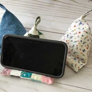 May include: A black smartphone rests on a small, colorful patchwork pillow. Two fabric wedges, one blue with white dots and the other white with a floral pattern, flank the phone. The items are arranged on a light wood surface.