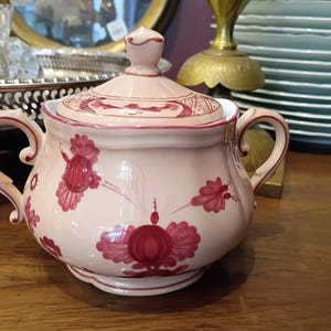 May include: A light pink ceramic sugar bowl with a matching lid. The bowl features two handles and is decorated with red floral designs. The lid has a finial and a similar floral pattern. The sugar bowl is on a wooden surface.