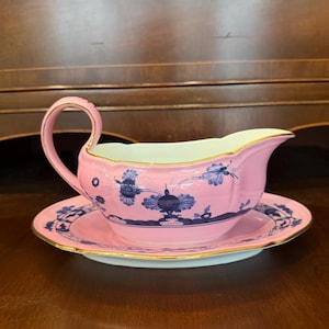 May include: Pink porcelain gravy boat and saucer set with blue floral designs and gold trim. The gravy boat has a curved handle and a spout. The saucer is round and complements the boat's design. A classic addition to any table setting.