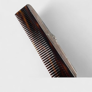 May include: A vintage comb with a tortoiseshell pattern and a silver metal backing. The comb's teeth are dark brown, and the silver backing has an ornate design. The comb is angled against a white background, showcasing its elegant design.