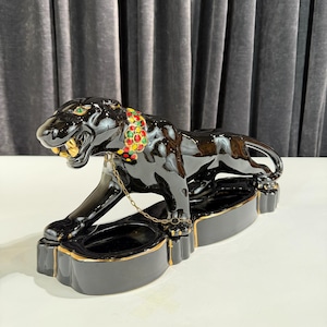 May include: A black ceramic panther figurine with gold accents. The panther has green eyes, a colorful jeweled collar, and a gold chain. The figurine is resting on a black base with a gold trim.