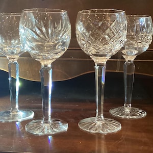 May include: Four clear crystal wine glasses with intricate cut patterns. The glasses have long stems and round bases. The cut designs include starburst and diamond patterns. The glasses are displayed on a wooden surface.