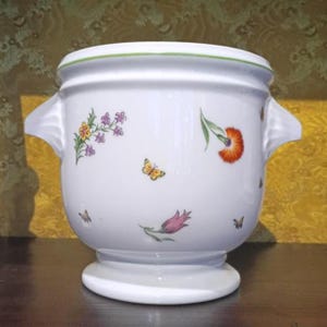 May include: A white ceramic cachepot featuring a flared rim with a thin green line, two side handles, and a pedestal base. The exterior is decorated with colorful illustrations of various flowers, including a pink tulip and orange bloom, and butterflies.